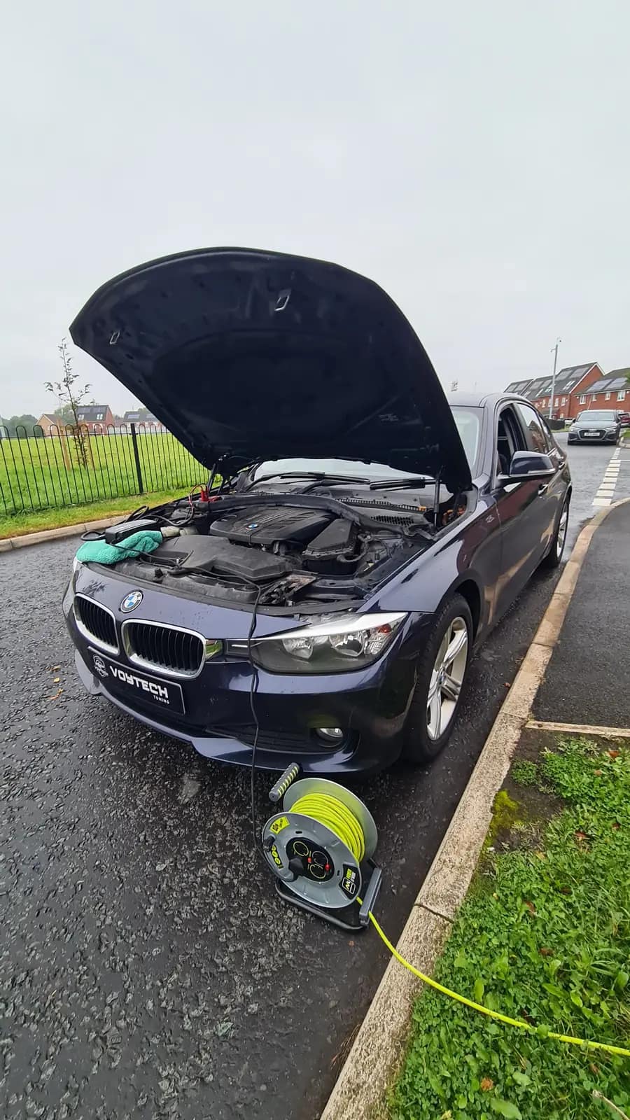 Dark BMW with the bonnet up and the kit connected on the street